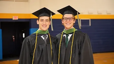 Two graduates smiling in graduation caps and gowns in the gym.